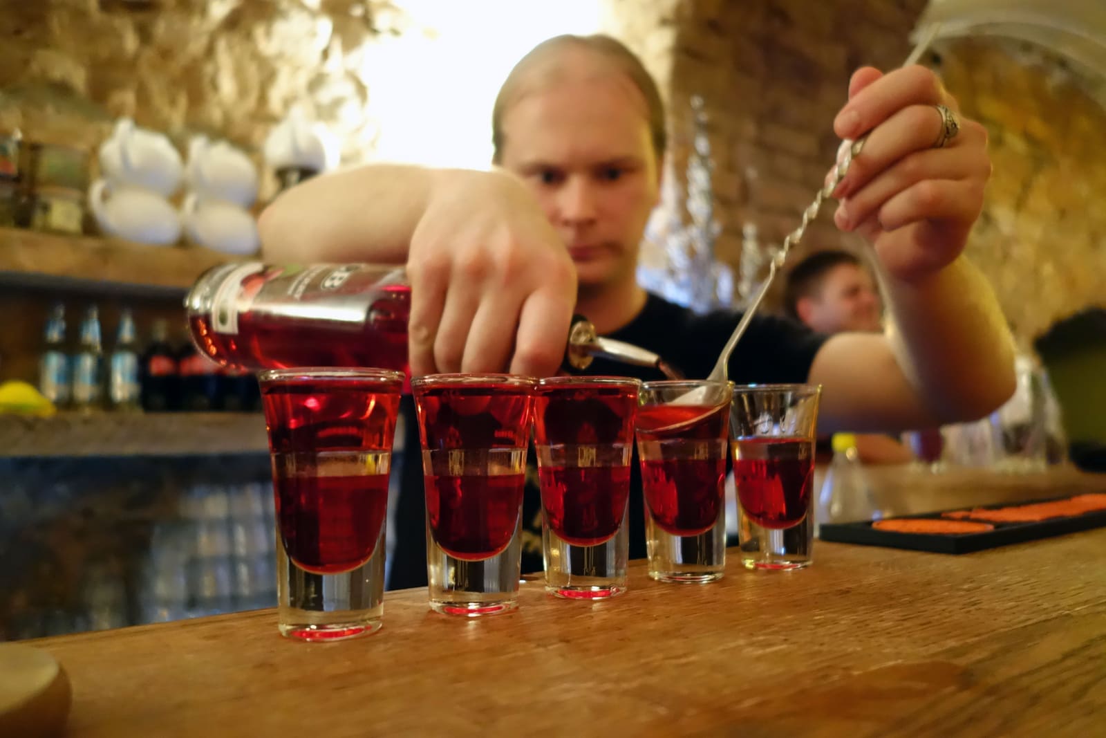 Bartender schenkt rode drank in shotglaasjes aan een houten bar met warm licht.