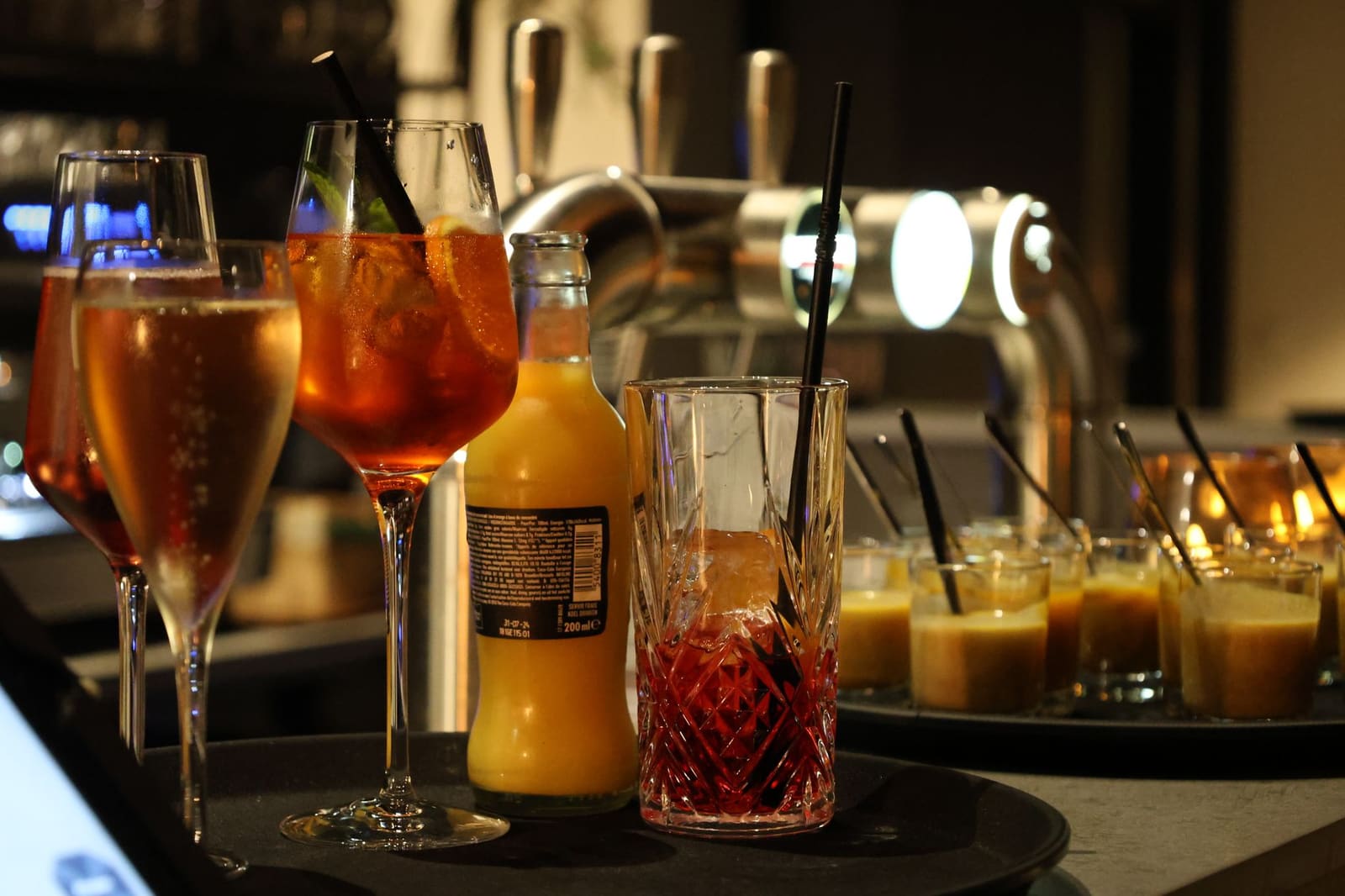 Verschillende cocktails en een flesje drank op een dienblad in een bar.