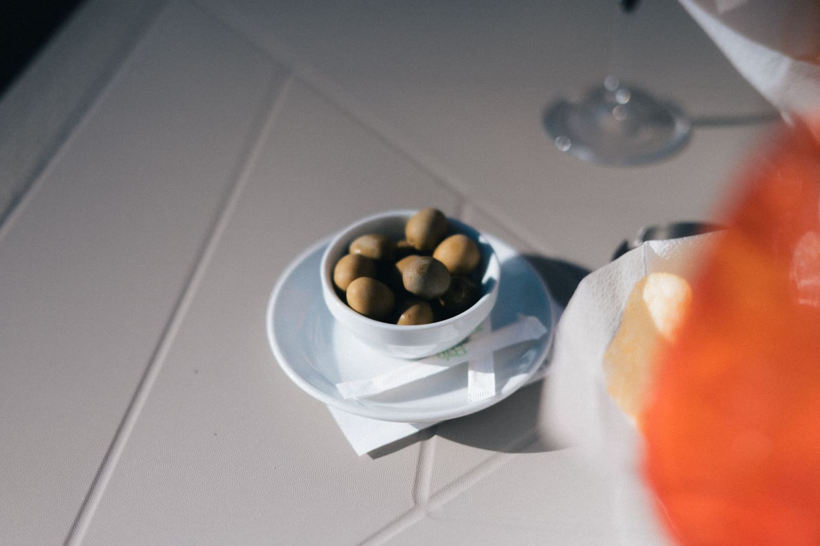 Schaaltje met olijven en chips naast een glas oranje drank op een terrastafel.