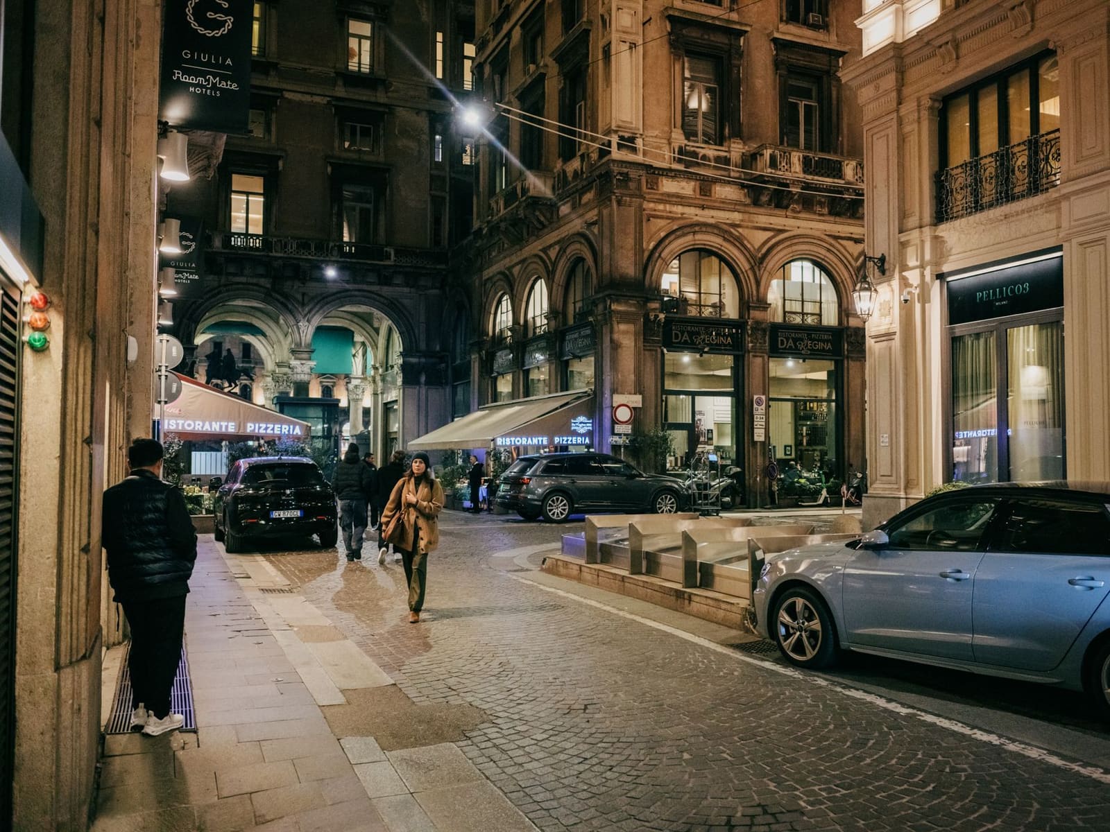 Verlichte Italiaanse straat 's avonds met restaurants, geparkeerde auto's en voorbijgangers.