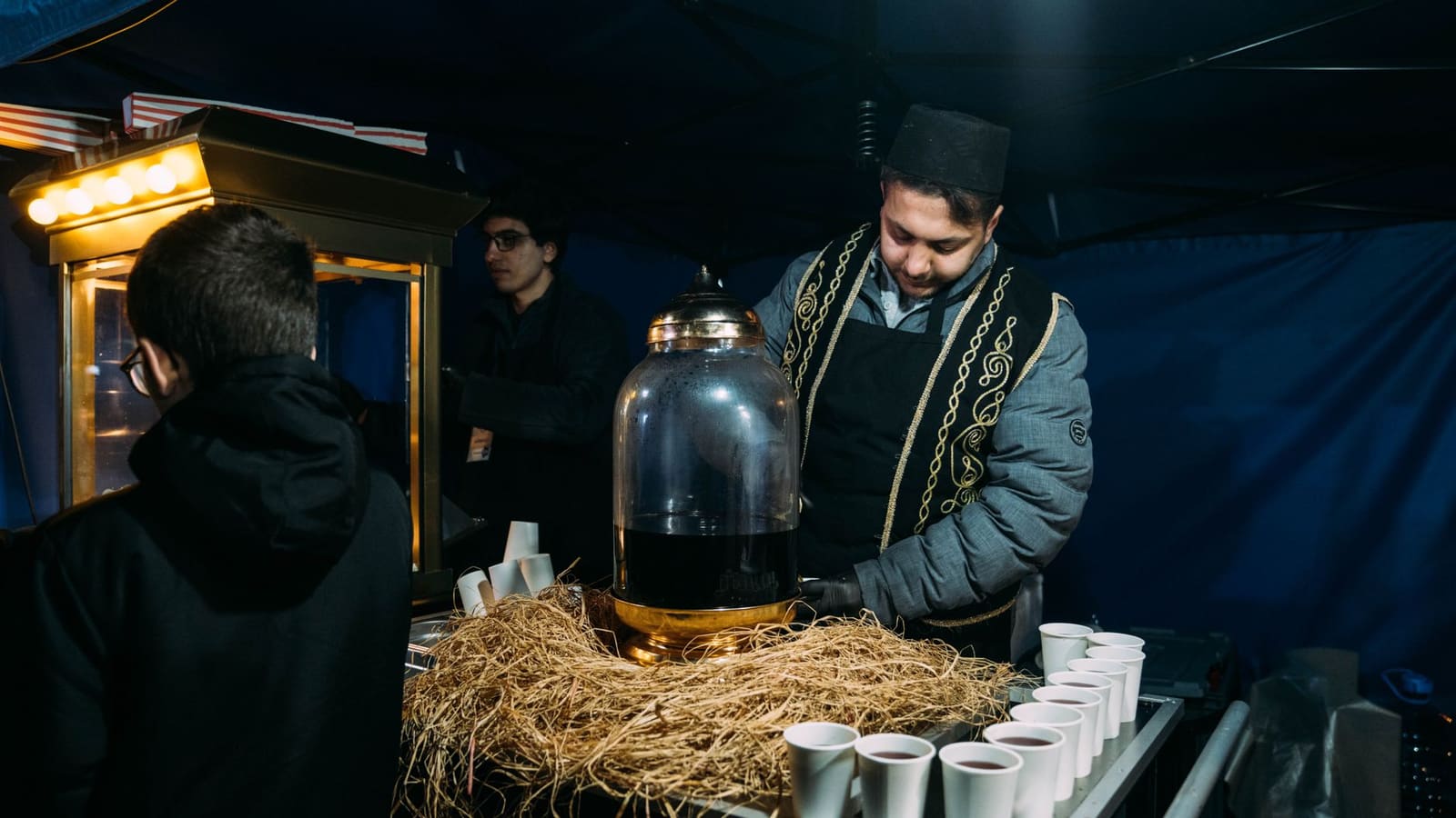 Een man in traditionele kledij schenkt drankjes uit een grote kan bij een kraam.