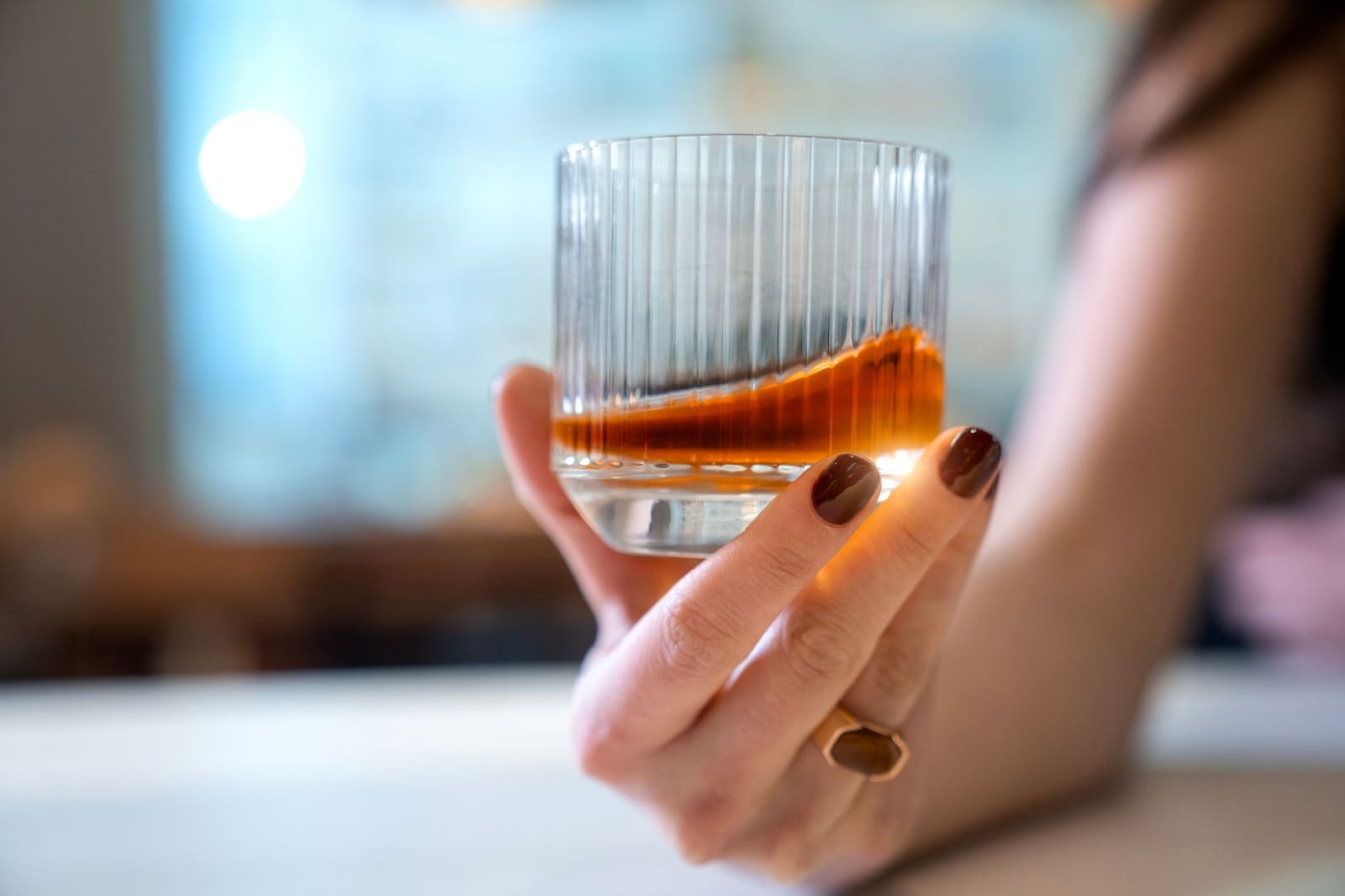 Hand met ring houdt geribbeld glas met amberkleurige drank vast, wazige achtergrond.