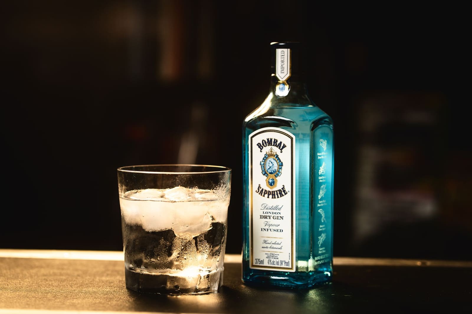 Fles Bombay Sapphire gin naast glas met ijsblokjes op bar.