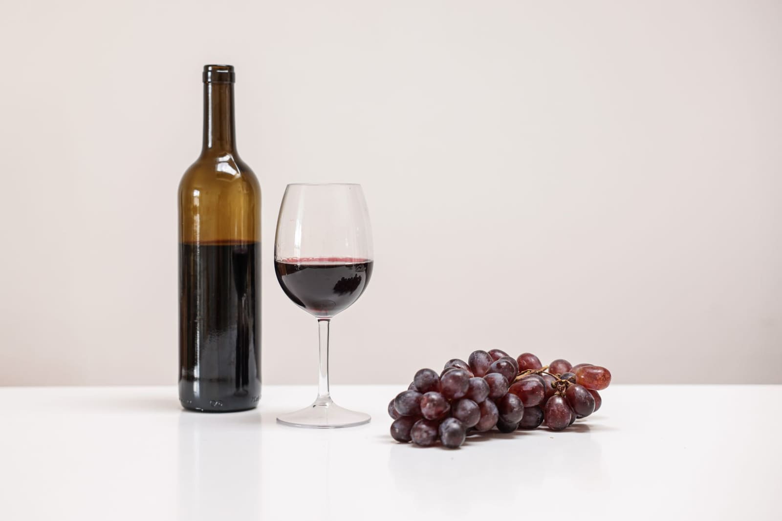 Glas rode wijn naast fles en druiventros op een witte tafel, neutrale achtergrond.