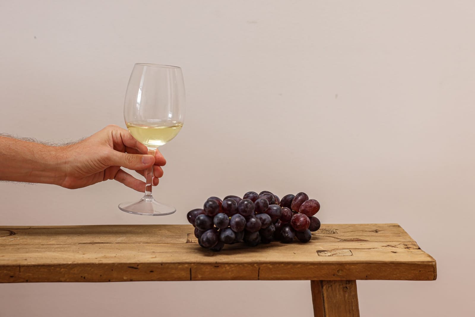 Hand houdt glas witte wijn vast naast druiventros op houten tafel