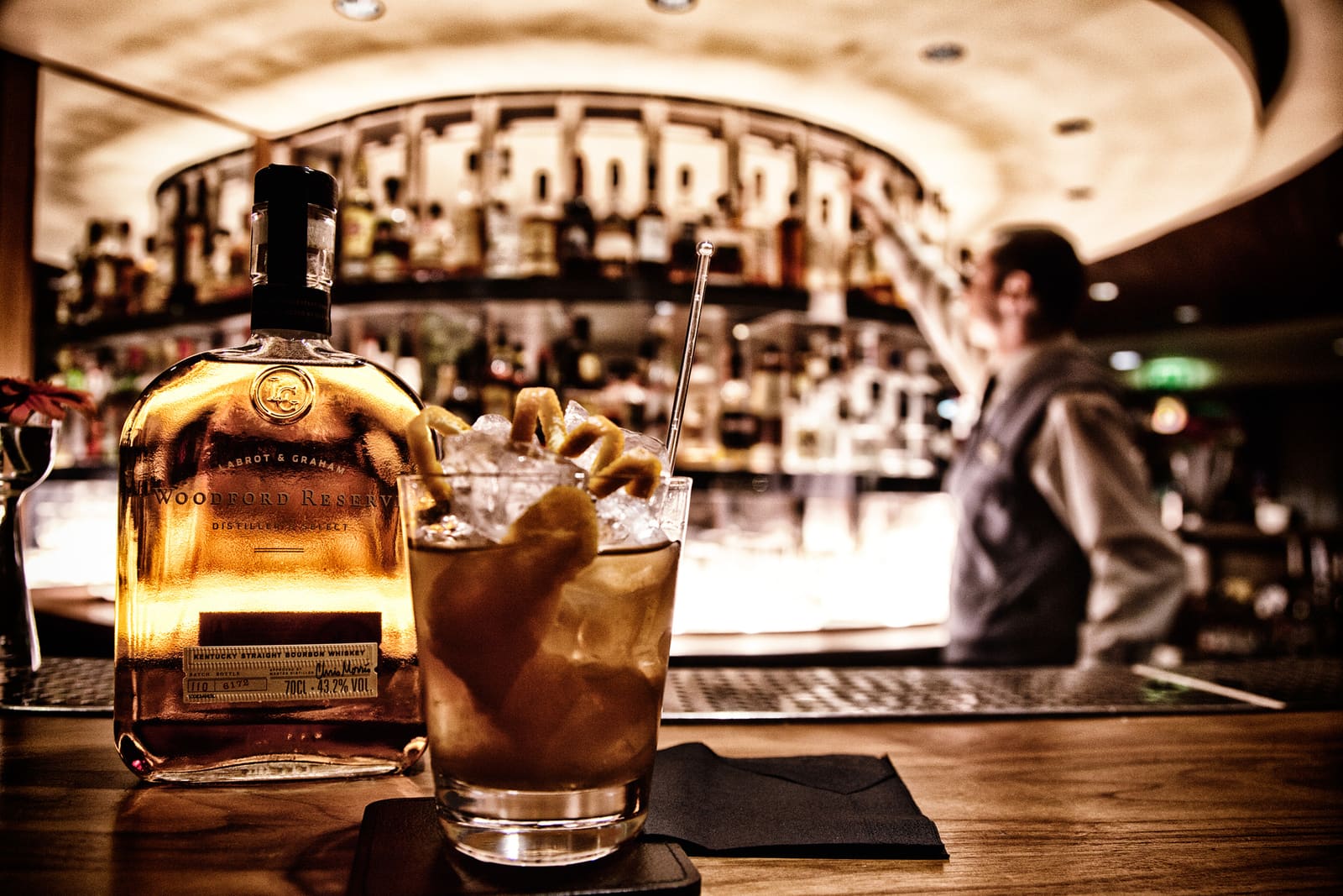 Een Old Fashioned met een grote ijsblok en een schil sinaasappel op een houten bar.