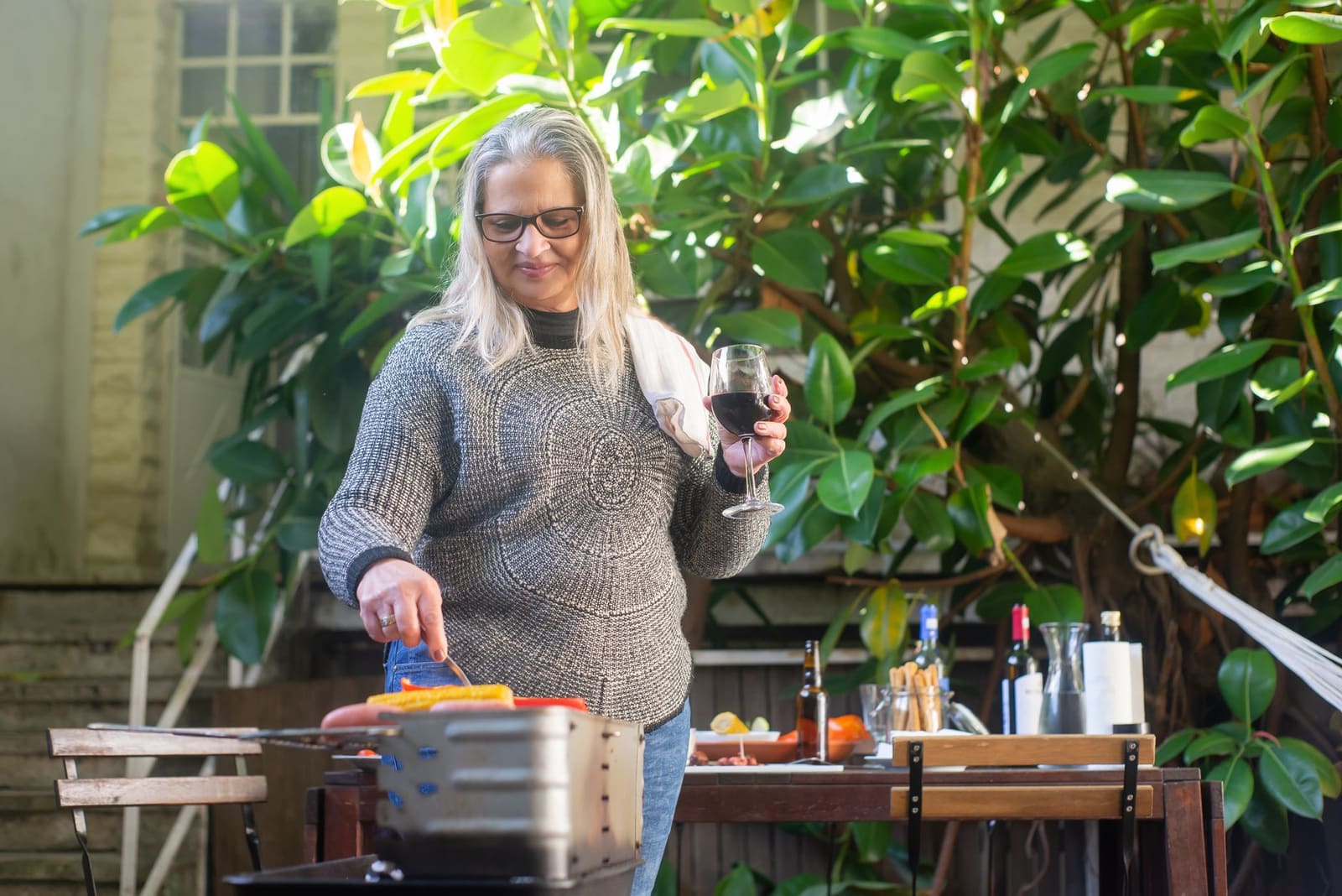 Vrouw bij tafelbarbecue houdt glas rode wijn vast tussen groene planten.