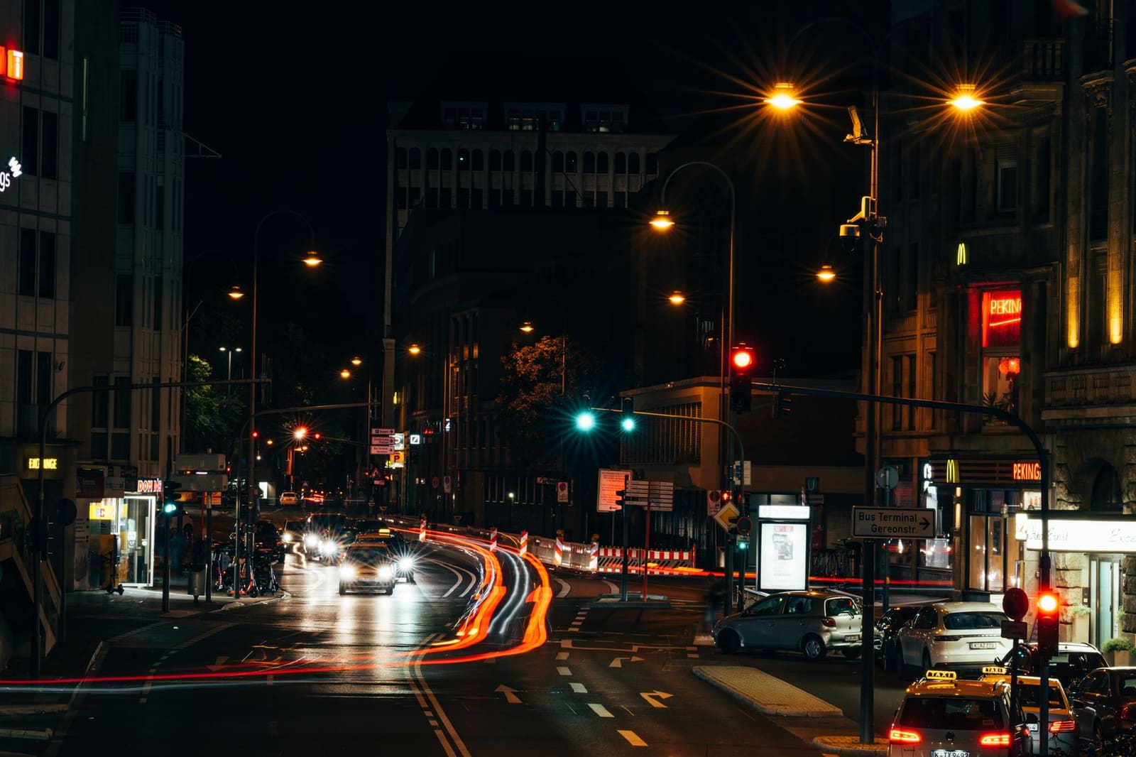 Verlichte stadsstraat bij avond met mensen die langs terrassen lopen.