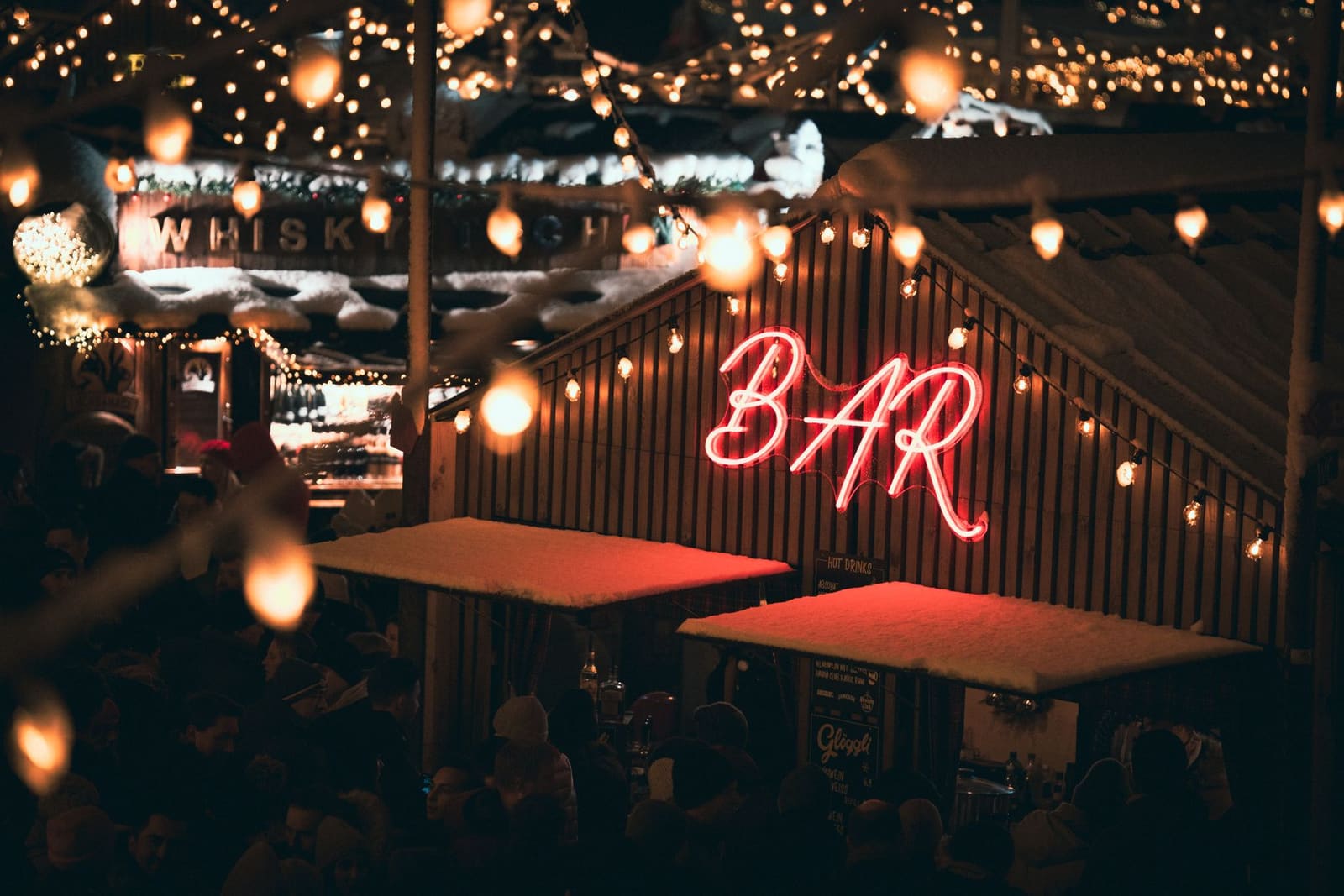 Buitenbar bij avond met neonbord 'BAR', lichtjes, sneeuw en mensen.
