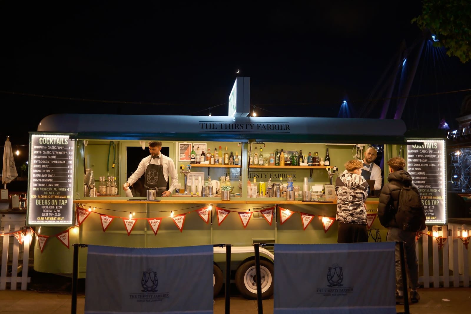 Barmedewerkers bereiden drankjes bij een verlichte mobiele bar op een festival 's avonds.