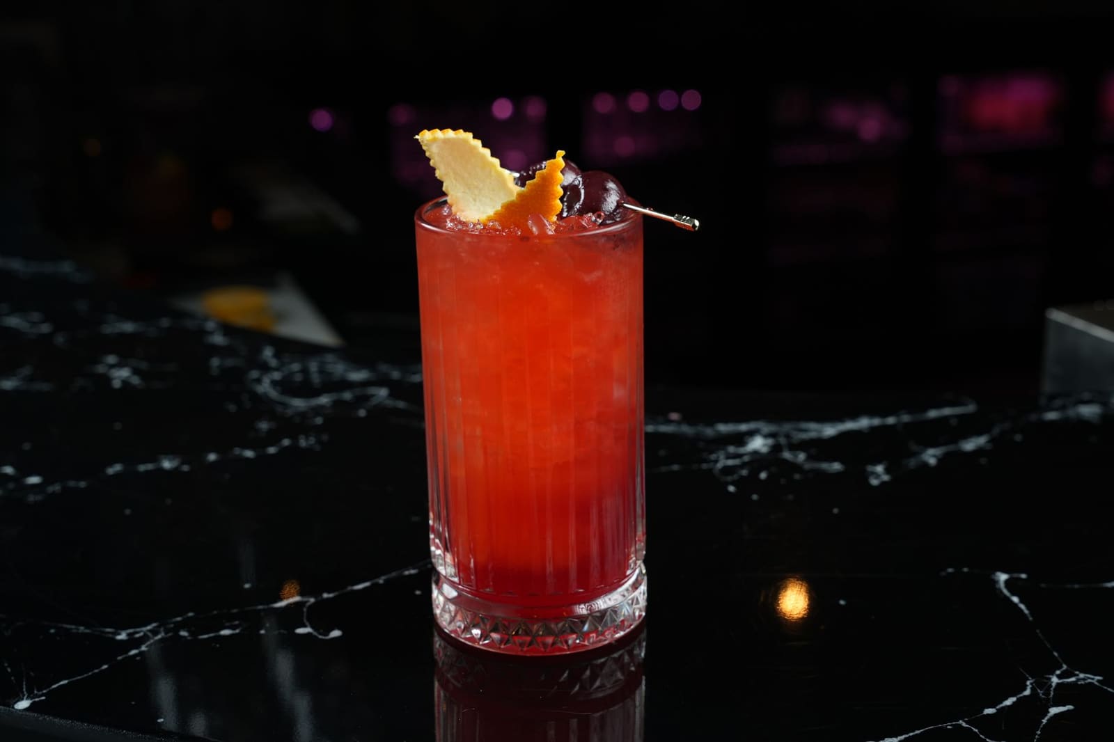 Rode cocktail met ijs, kersen en citrusschil garnering op donkere bar.