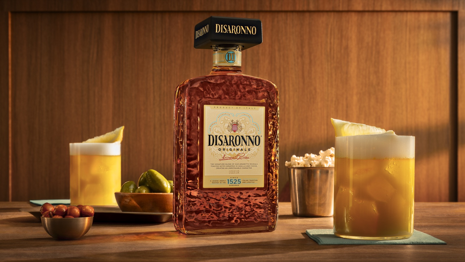 De vernieuwde Disaronno-fles met zwarte vierkante dop tussen twee Disaronno Sours op een houten bar