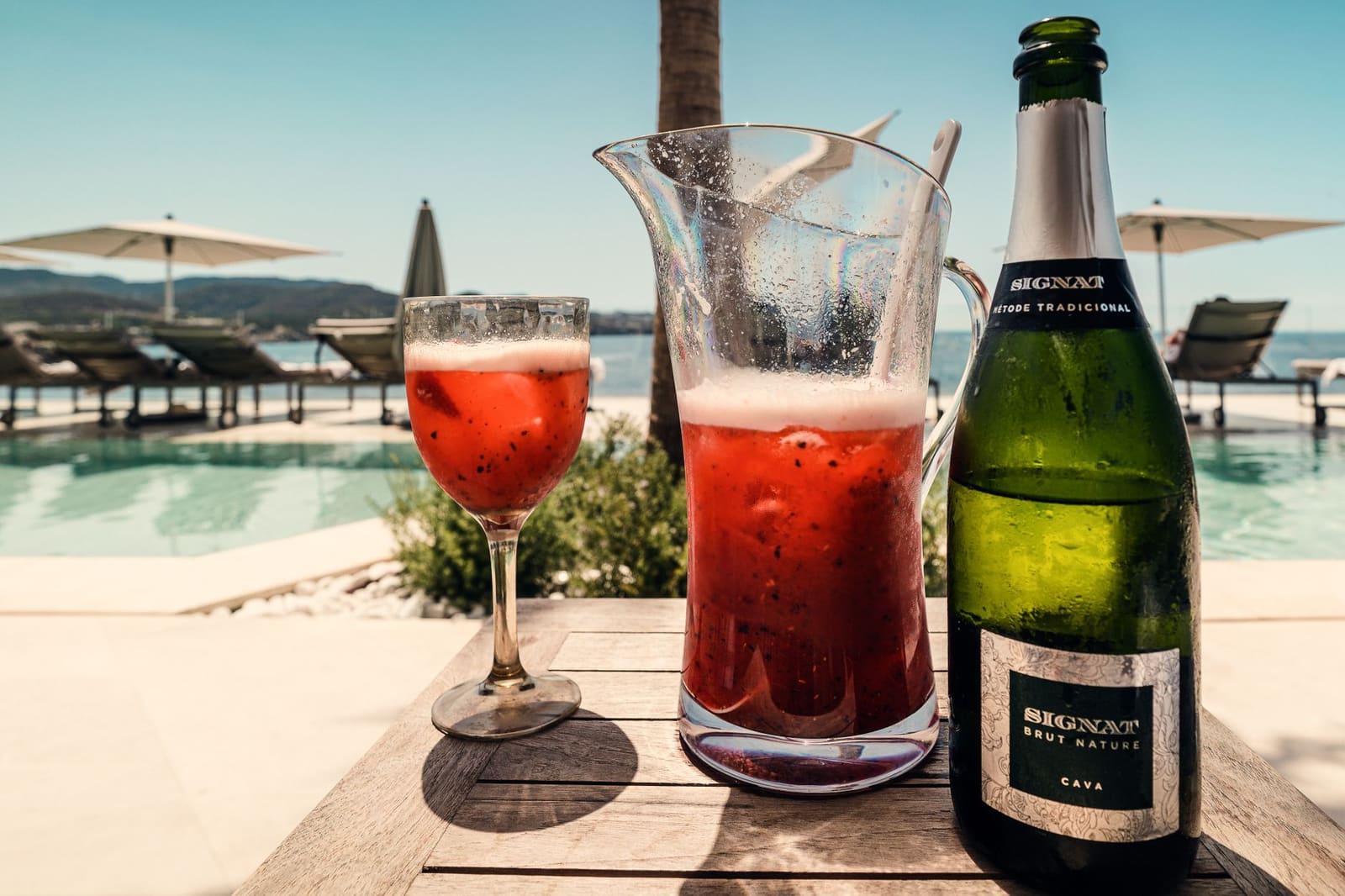 Glas en kan met rode cocktail en fles cava bij zwembad aan zee
