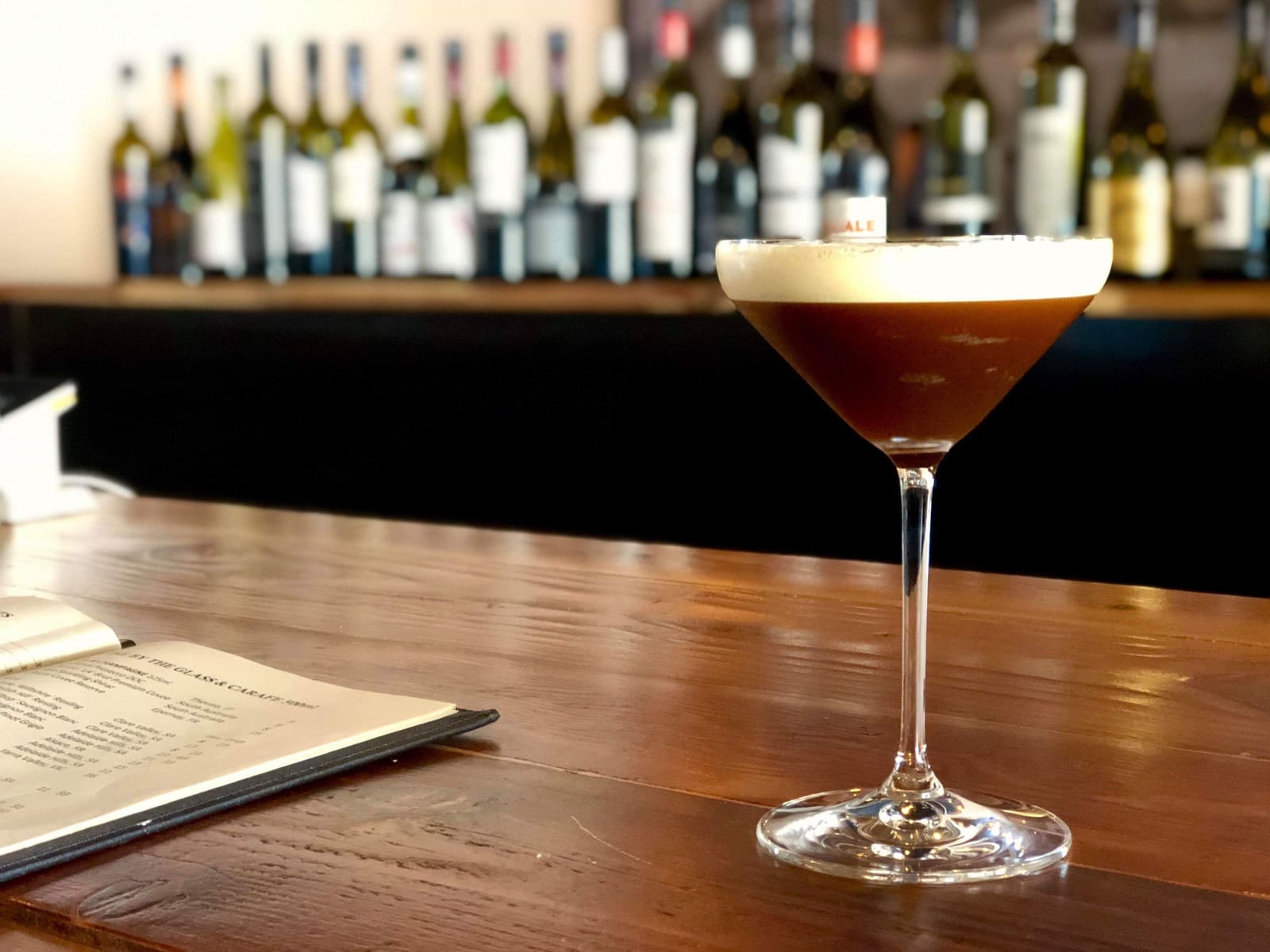 Espresso martini met schuim op de bar, achtergrond met wijnflessen en menukaart.