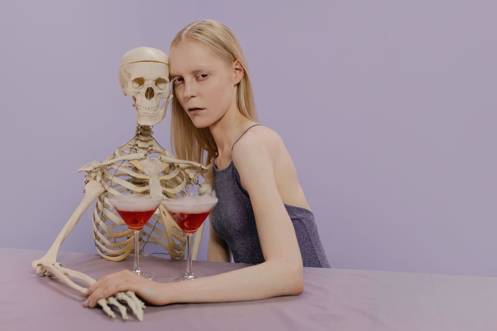 Blonde vrouw naast skelet drinkt rode cocktail met rook aan tafel