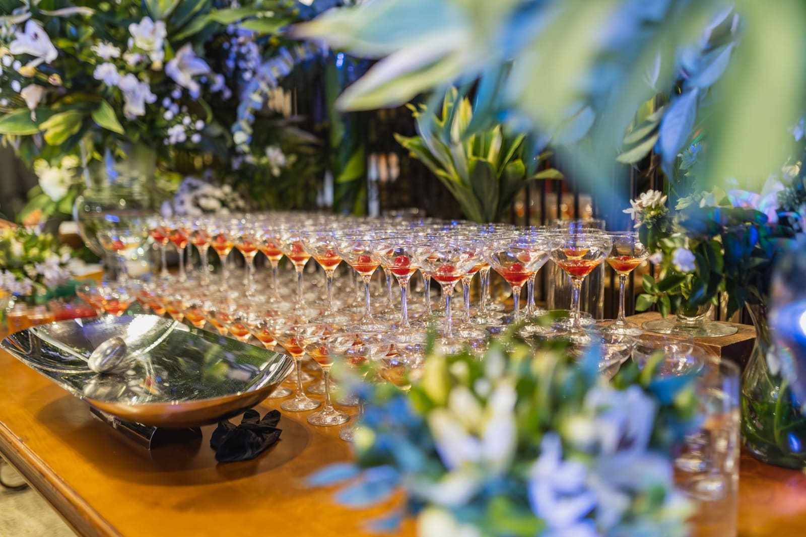 Rij cocktailglazen met rode drankjes en kers, feestelijk gedekte tafel met bloemen.