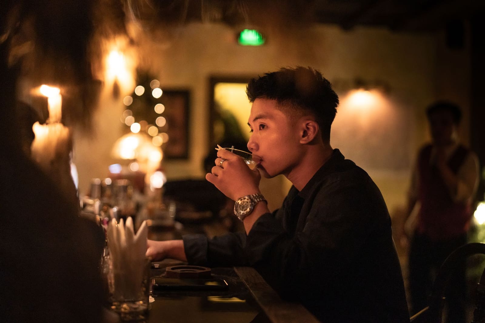 Man drinkt een cocktail aan een donkere bar met kaarslicht en wazige lichtjes op achtergrond.