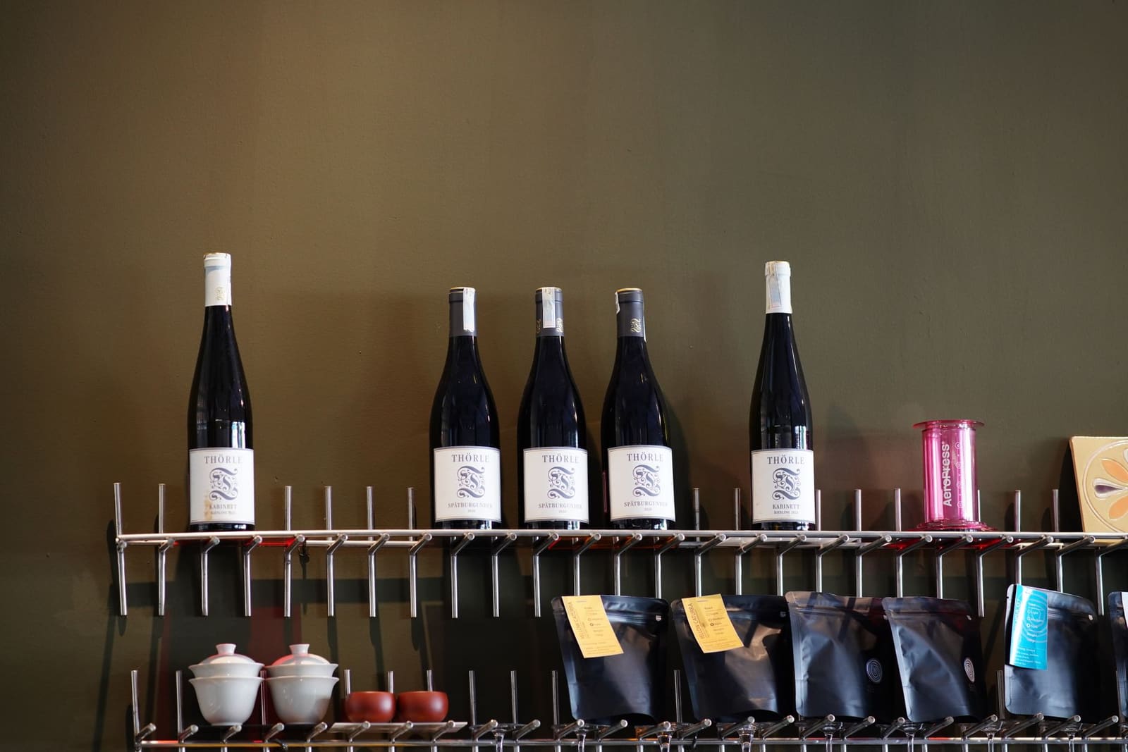 Een plank met wijnflessen, koffiezakjes, theekopjes en een AeroPress in een winkel.