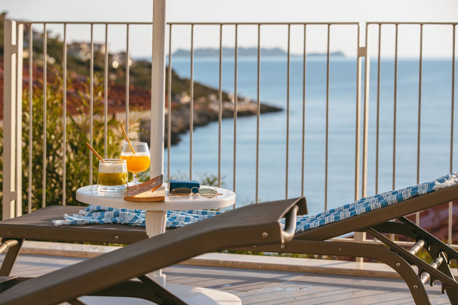 Twee cocktails met rietjes op terrastafel bij zee tijdens zonsondergang, loungestoelen op voorgrond.