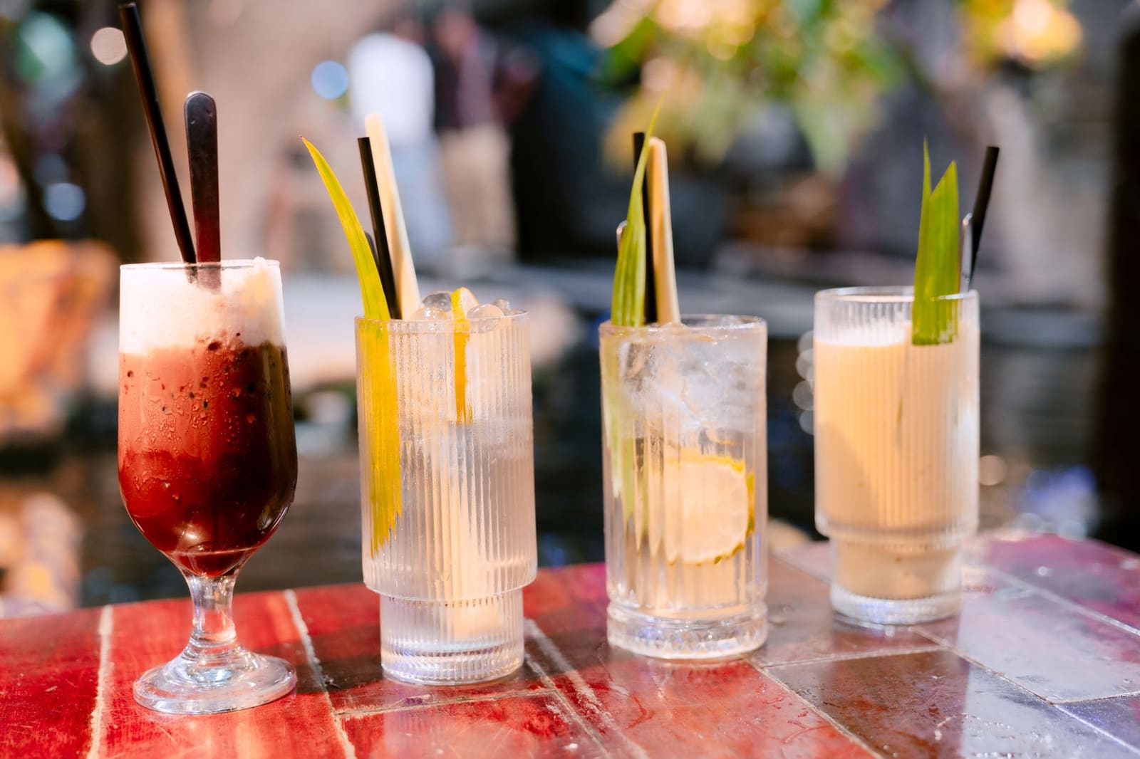 Vier cocktails met rietjes en bladgarnering op rood-houten tafel, vage achtergrond.