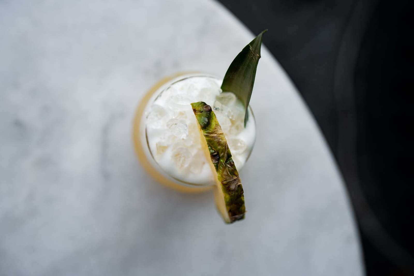 Bovenaanzicht van witte cocktail met crushed ice en ananasschijf op marmeren tafel.