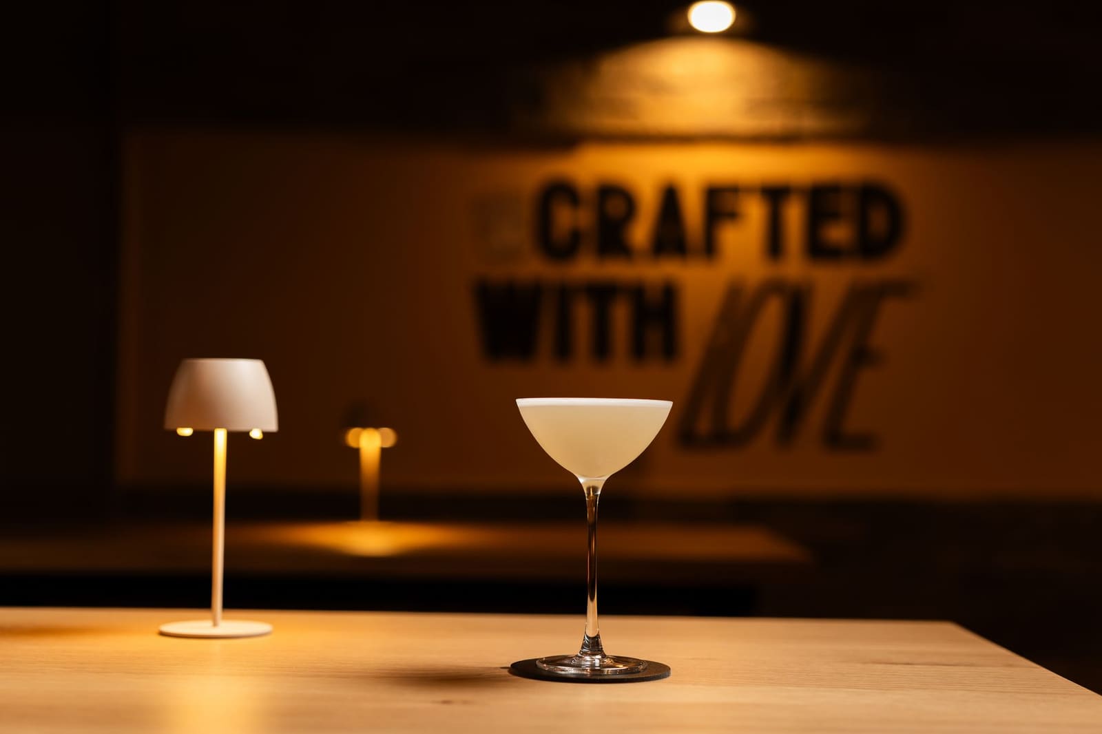 Lichtgele cocktail met schuimlaag in coupeglas op houten bar, warme sfeerverlichting op achtergrond.