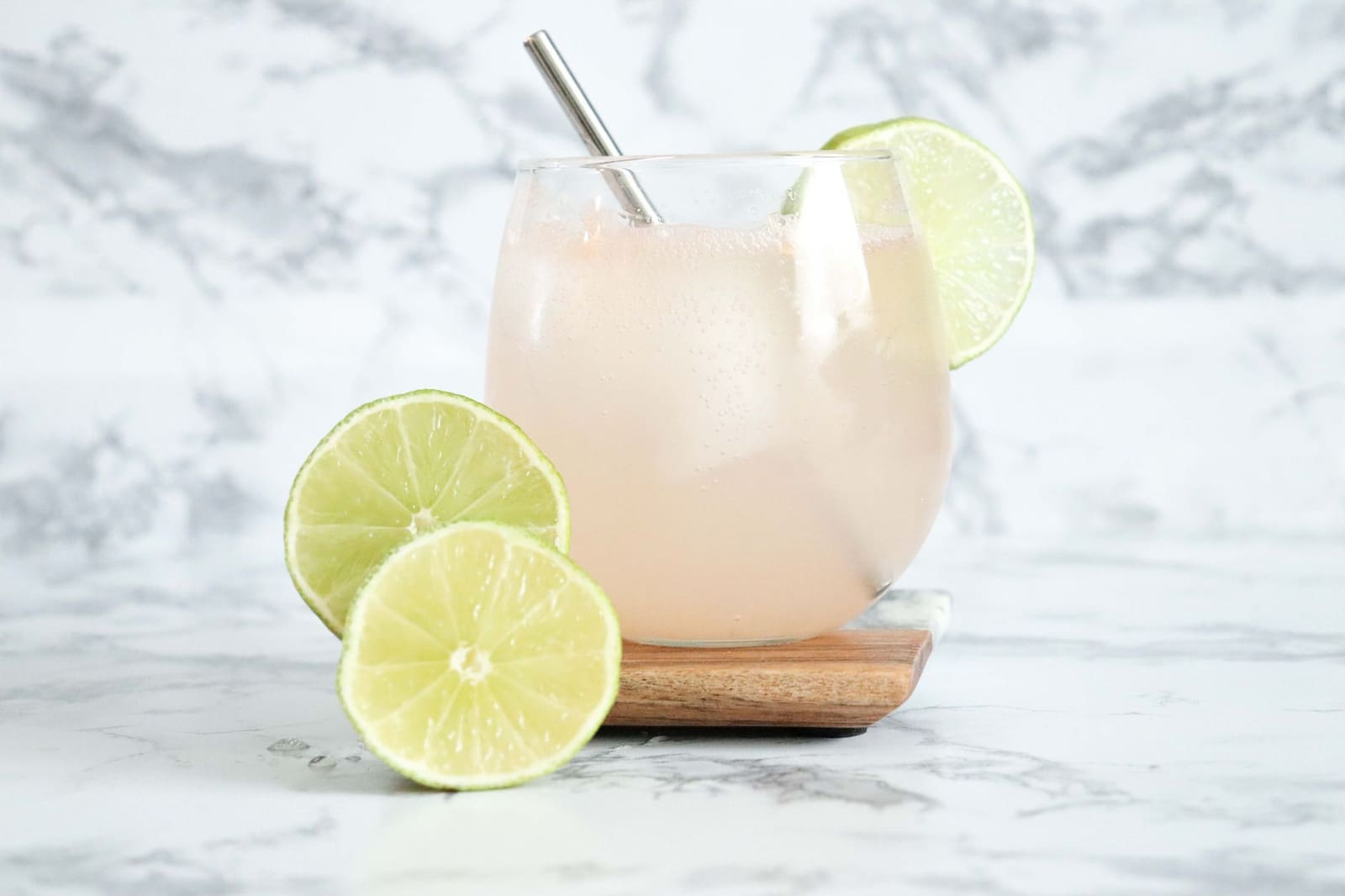 Glas met lichtroze drank, ijs, rietje en limoenschijfjes op marmeren ondergrond.