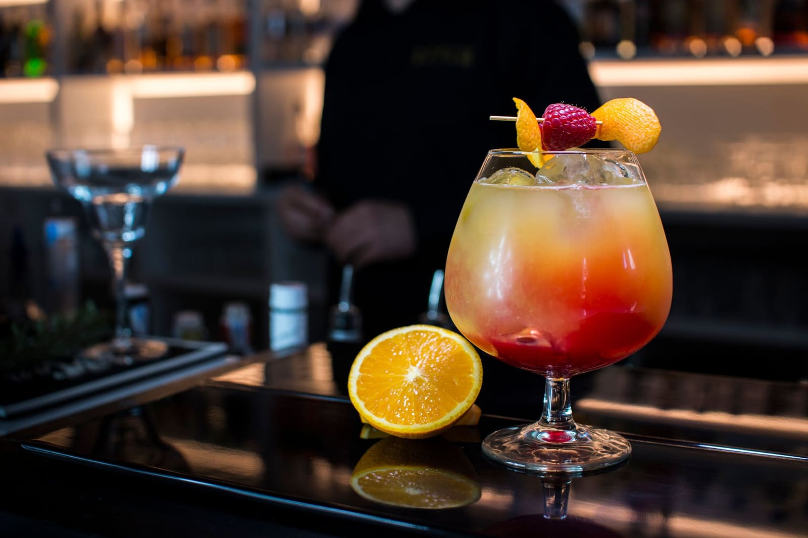 Tequila Sunrise met sinaasappel en rode laag, gegarneerd met framboos, geserveerd aan bar