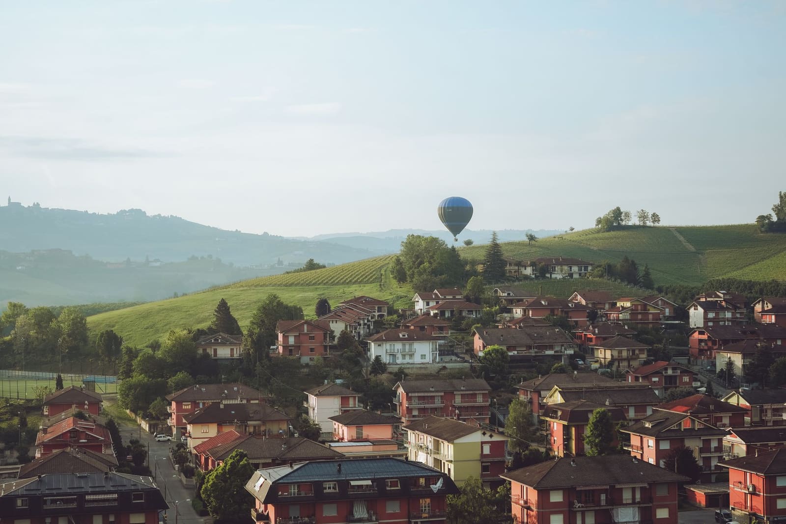 Luchtfoto van dorp met rode daken, heuvels met wijngaarden en een heteluchtballon.