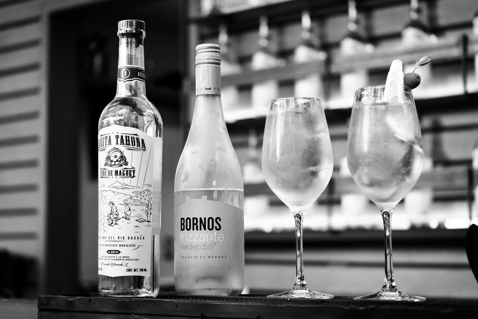 Flessen mezcal en wijn met twee glazen cocktail op een bar, zwart-wit foto.